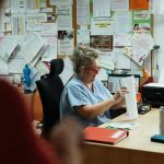 A senior caregiver in blue scrubs sorting paperwork at a hospital office desk.